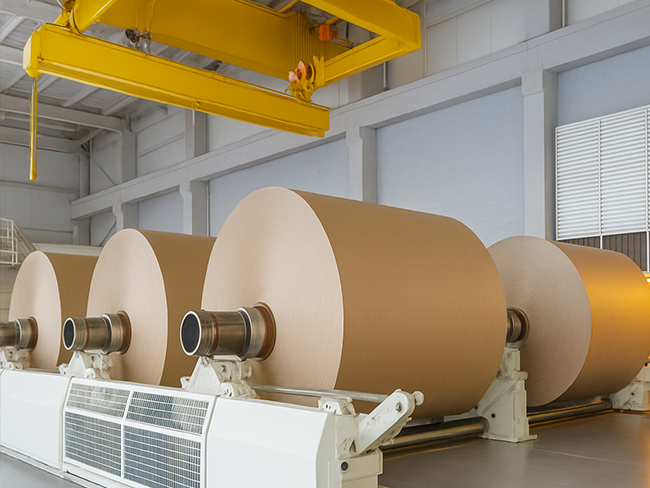 Massive rolls of Paktek paper mounted on industrial winding machinery in a bright factory.